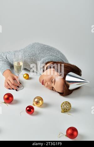 Frau in Partymütze schläft auf einem Tisch neben Sektglas und Weihnachtskugeln vor grauem Hintergrund Stockfoto