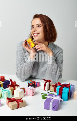 Fröhliche Frau im Pullover, die ein Weihnachtsgeschenk in der Nähe von Geschenken auf dem Tisch hält Stockfoto