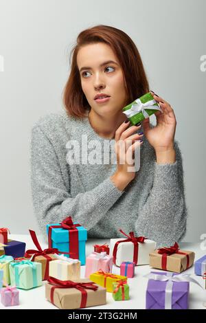 Schöne Frau in einem gemütlichen Pullover, der ein kleines Weihnachtsgeschenk in der Nähe von bunten gewickelten Geschenken auf Grau hält Stockfoto