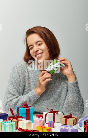 Fröhliche Frau in einem gemütlichen Pullover, der ein kleines Weihnachtsgeschenk in der Nähe von bunten gewickelten Geschenken auf Grau hält Stockfoto