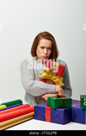 Verärgerte Frau im Winterpullover mit rotem umwickeltem Weihnachtsgeschenk in der Nähe von Geschenkpapier auf grauem Hintergrund Stockfoto
