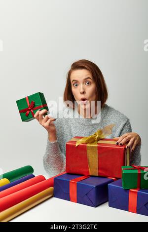wow Emotion, Frau im Winterpullover mit Weihnachtsgeschenk neben farbenfrohem Geschenkpapier auf Grau Stockfoto