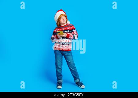 Foto in voller Länge von hübschen träumerischen kleinen Jungen tragen Ornament Pullover Xmas Hut chatten Gadget leeren Raum isolierten blauen Farbe Hintergrund Stockfoto