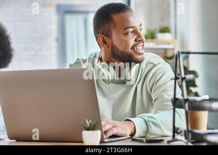 Fröhlicher Mann im urbanen Outfit mit Zahnspangen, die wegschauen, während sie mit Laptop arbeiten, Business-Konzept Stockfoto