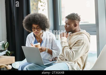 Konzentrierte Freunde in lässigen Outfits mit Kaffeetasse in Händen, die an einem Laptop arbeiten, Business-Konzept Stockfoto