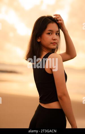 Ein asiatischer Teenager mit einem süßen Gesicht in schwarzer Kleidung lächelt und genießt die wunderschöne Landschaft am Strand vor Sonnenuntergang Stockfoto