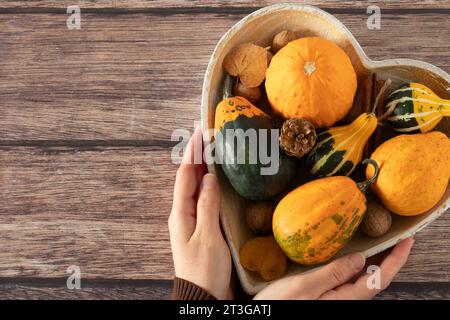 Weibliche Hände halten eine herzförmige Schüssel mit dekorativen Kürbissen, Nüssen und Herbstblättern auf Holztisch. Draufsicht, Kopierraum. Thanksgiving-Konzept. Stockfoto