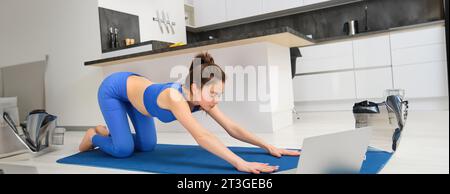 Porträt einer Frau, die Dehnübungen macht, folgt dem Tutorial auf dem Laptop, nimmt an Online-Yogastunden von zu Hause aus Teil, trainiert auf einer Gummimatte Stockfoto