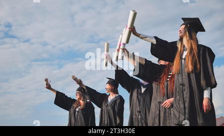 Fröhliche Absolventen posieren mit erhöhten Diplomen an einem sonnigen Tag. Stockfoto