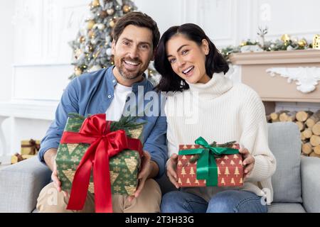 Porträt einer glücklichen jungen Familie, eines Mannes und einer Frau, die zu Hause auf dem Sofa sitzen, Geschenkboxen halten, lächeln und glücklich für die Kamera sind. Stockfoto