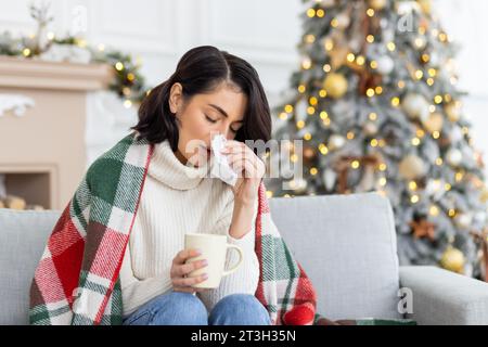 Eine junge Frau erkrankte während der Weihnachts- und Neujahrsferien. Sie sitzt zu Hause auf dem Sofa, das mit einer Decke bedeckt ist, wischt sich mit einer Serviette die Nase von einer laufenden Nase ab, hält eine Tasse Drink, Medizin. Stockfoto