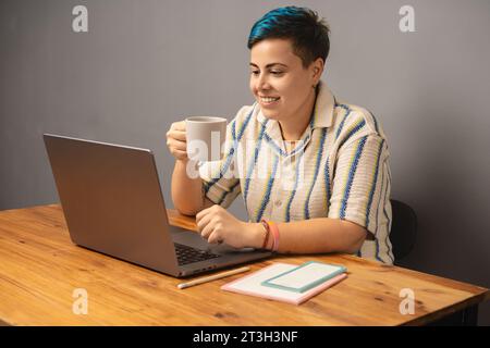 Eine moderne und stilvolle Frau mit blauem Haar sitzt in ihrem Home Office, schlürft Kaffee und arbeitet an ihrem Laptop Stockfoto