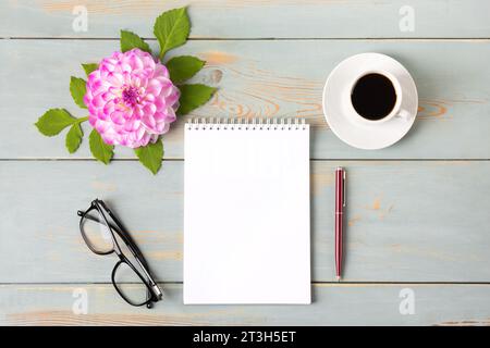 Draufsicht des Desktops. Spiralnotizbuch mit offener leerer Seite, Gläser, Stift, weiße Tasse mit schwarzem Kaffee, Dahlienblüten. Festliches Büro Desktop Co Stockfoto