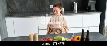 Porträt einer glücklichen jungen Frau schreibt Menü zum Abendessen auf, sitzt in der Küche neben Gemüse, macht Lebensmittellisten zum Einkaufen, posiert im Bademantel Stockfoto