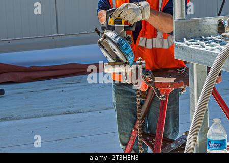 Ein Bauarbeiter schneidet mit einer tragbaren Bandsäge Edelstahlrohre auf dem Dach einer industriellen Kühlanlage (Kühllager). Stockfoto