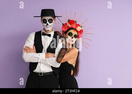 Junges Paar, das sich für den mexikanischen Tag der Toten (El Dia de Muertos) auf Fliederhintergrund verkleidet hat Stockfoto