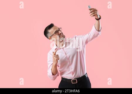 Glücklicher junger Geschäftsmann mit Handy, der Selfie auf rosa Hintergrund macht Stockfoto