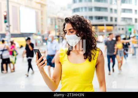 Frau mit Maske, die auf das Handy in der Stadt schaut Stockfoto