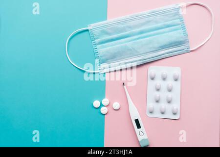 Schutz vor Coronavirus-Infektion: Eine blaue Einwegmaske, weiße Pillen, ein elektronisches Thermometer auf hellrosa und türkisfarbenem Hintergrund. Stockfoto