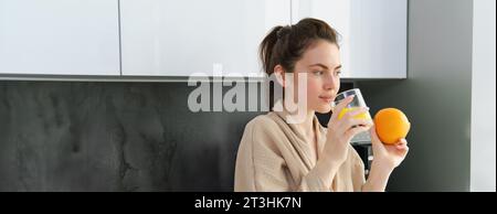 Junge Frau, die frisch gepressten, selbstgemachten Orangensaft in der weißen Küche trinkt Stockfoto