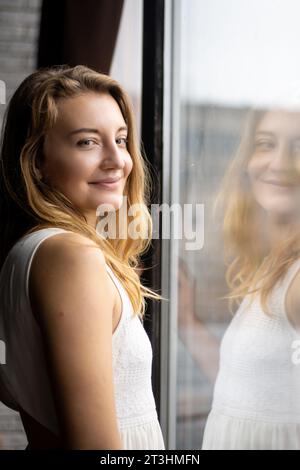 Eine junge Dame in einem fließenden weißen Kleid grinst, sich im sanften Licht des Sonnenlichts nahe einem großen Fenster zu erfreuen. Strahlende Positivität füllt den Raum und spiegelt den wider Stockfoto