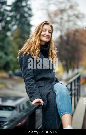 Eine Frau in einem langen grauen Mantel und einer blauen Jeans auf weißen Turnschuhen sitzt auf dem Geländer und schaut auf die Kamera auf der Straße einer Herbststadt Stockfoto
