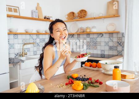 Eine junge, attraktive asiatische Frau isst frische Beeren in der Küche. Ein charmantes Mädchen hält einen Teller mit Himbeeren, Erdbeeren und Heidelbeeren und Hols Stockfoto