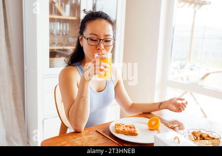 Eine lächelnde, attraktive asiatische Frau trinkt frisch gepressten Orangensaft. Eine charmante junge Koreanerin hält ein Glas köstlichen Saft und lacht in der Th Stockfoto