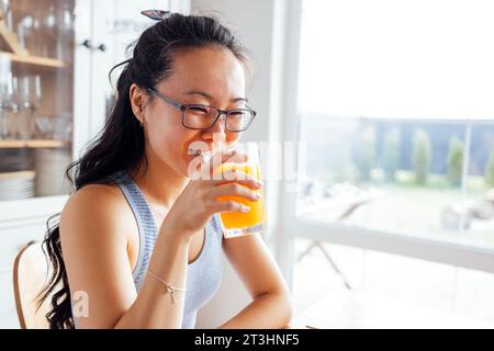 Eine lächelnde, attraktive asiatische Frau trinkt frisch gepressten Orangensaft. Eine charmante junge Koreanerin hält ein Glas köstlichen Saft und lacht in der Th Stockfoto