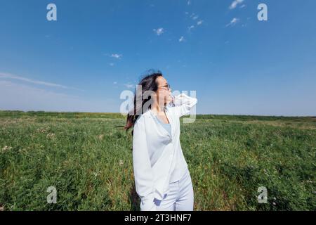 Eine süße junge asiatische Frau mit Brille lächelt vor einem Hintergrund aus blauem Himmel und grünem dichtem Gras. Ein charmantes koreanisches Mädchen in lässiger Kleidung ist entspannt Stockfoto