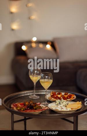 Zwei Gläser kühlen Sekt und Teller mit verschiedenen Snacks auf einem runden Metall-Couchtisch. Weiße Teller mit Schinkenscheiben, Salami, Kapern, Oliven und Stockfoto