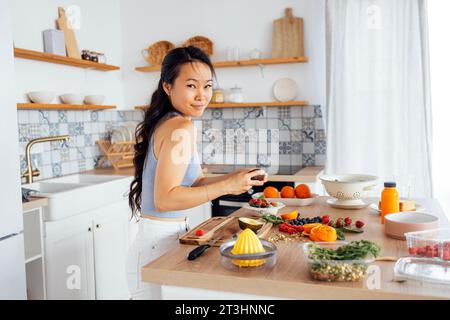 Eine attraktive süße asiatische Frau lächelt und kocht das Frühstück in der Küche. Eine junge charmante Koreanerin lacht und schneidet Gemüse und Fisch. Gesund f Stockfoto