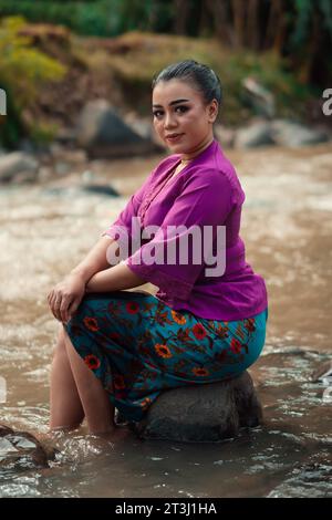 Schöne asiatische Frau, die auf dem Felsen in der Nähe des Flusses sitzt und ein Lächeln trägt, während sie im Dorf ein lila Kleid trägt Stockfoto