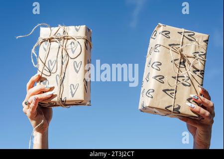 Foto von der ersten Person von oben mit weiblichen Händen, die zwei Geschenkboxen aus Papier über blauem Himmel halten Stockfoto