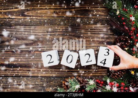 Frauenhand kippt die 2023 Holzkalenderblöcke des Neujahrs auf 2024. Weihnachtsbaumbeleuchtung, Tannenzweige, rote Winterbeeren und Schnee. Draufsicht. Stockfoto