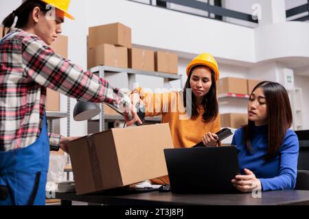 Asiatische Paketabwickler und Zusteller bereiten das Paket für den Versand vor. Mitarbeiter des Postlagers verwenden Barcode-Scanner und überprüfen Kundenbestellungen auf einem Laptop Stockfoto