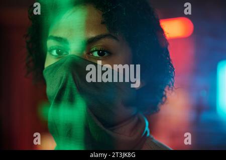 Gesicht des afroamerikanischen Mädchens halb mit Maske bedeckt und in die Kamera schaut, während er in der Cyberpunk Bar mit Neonlichtern auf dem Hintergrund steht Stockfoto