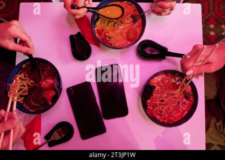 Blick von oben auf mehrere Schüsseln mit appetitlichen japanischen Ramen und Händen von Freunden mit Essstäbchen, die Essen am Tisch mit zwei Smartphones essen Stockfoto