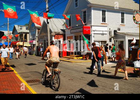 Die Leute laufen und fahren mit dem Fahrrad entlang der Commercial Street, der Hauptstraße in Provincetown, Cape Cod, mit portugiesischen Fahnen, die an den Gebäuden hängen Stockfoto