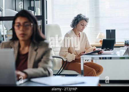 Ernsthafte, reife Direktorin des Unternehmens liest Geschäftsdokumente und überprüft deren Punkte, während sie vor dem Untergebenen am Schreibtisch sitzt Stockfoto