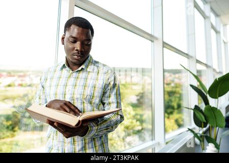 Afrikanischer Mann, der neben dem Fenster im Zimmer steht und ein Buch liest Stockfoto