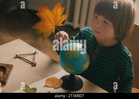 Das freimütige, verspielte Porträt eines blonden Jungen am Tisch, der mit Globus und Herbstlaub lernt und spielt Stockfoto