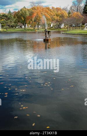 Ein dunkler Teich mit treibenden Blättern liegt vor einer kleinen Brunnenstatue und Herbstbäumen am fernen Ufer auf dem Forest Lawn Cemetery in Buffalo, New York. Stockfoto