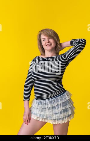 Fröhliche Blondine 49 Jahre in Minirock und gestreiftem T-Shirt posiert mit Hand hinter Kopf. Model lächelt und blickt weg. Porträt des fröhlichen W Stockfoto