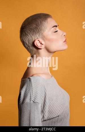 Profil der Seitenansicht einer nachdenklichen jungen blonden Frau mit kurzen Haaren und geschlossenen Augen, die im Raum mit dem Kinn nach oben gegen die orangene Wand steht Stockfoto