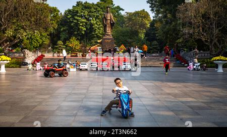 Ein junger vietnamesischer Junge spielt auf einem Dreirad vor der riesigen Bronzestatue des legendären Kaisers Ly Thai To im Zentrum von Hanoi Stockfoto