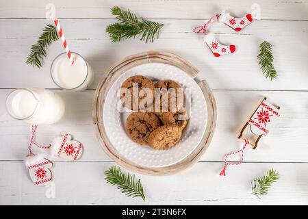 Weihnachtsarrangement mit Glasmilch, Keksen, Weihnachtssymbolen Weihnachtsschlitten, Weihnachtsmännern, Weihnachtsmännern Socken und kleinen Tannenzweigen auf weißem Holztisch Stockfoto