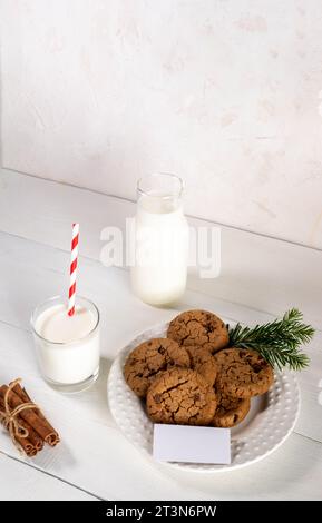 Frische Plätzchen auf rundem Teller, Milch in Glas und Flasche, leere leere leere Notiz auf weißem Holztisch vor weißer Wand. Weihnachtstradition - Milch für Sant Stockfoto