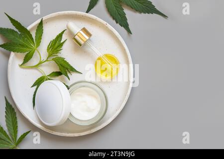 Geöffnetes Reisgefäß mit leerem, weißem Deckel und Pipette mit Öl in der Nähe von grünen Cannabis Sativa Blättern auf Teller auf grauer Tischansicht. Bio-Hautpflege Beauty p Stockfoto