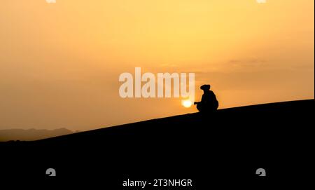 Silhouette eines nicht identifizierten Berbermanns, der den Sonnenuntergang auf Sanddünen bei einem wunderschönen Sonnenuntergang in der Sahara, Marokko, beobachtet Stockfoto